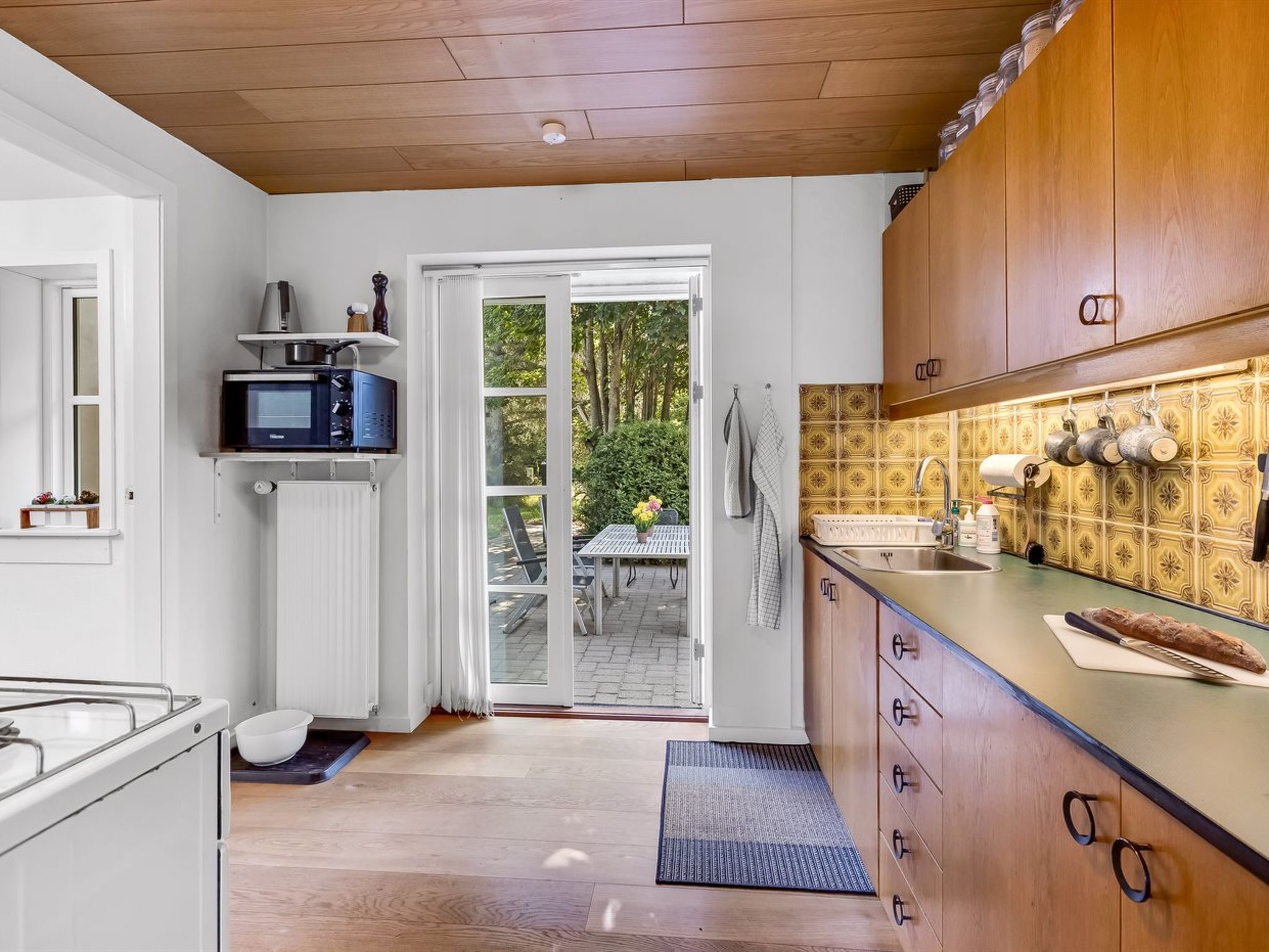 Una cocina con armarios blancos, un microondas y un lavamanos. Hay una puerta que da a un espacio exterior con una mesa y sillas.