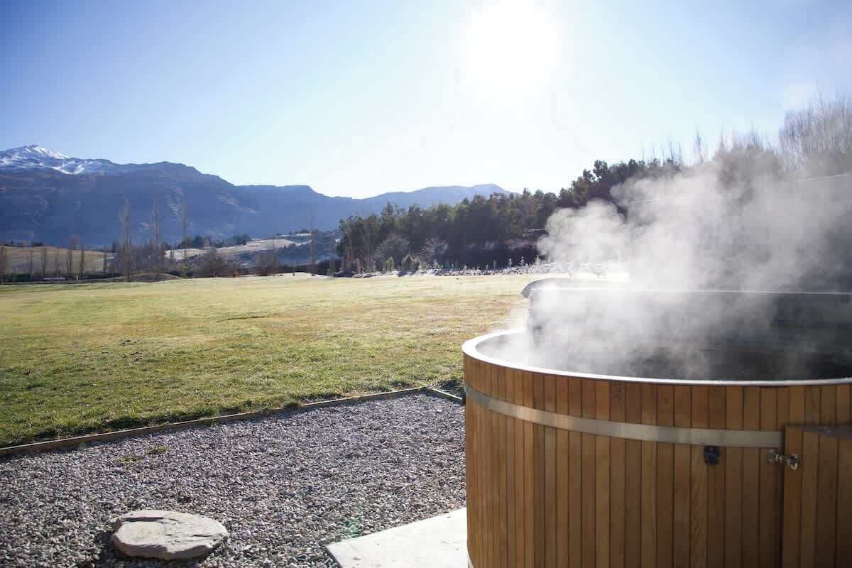 Un sauna en bois d’où s’échappe de la vapeur, se dressant sur un fond de prairie verdoyante et de montagnes lointaines.