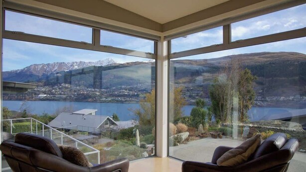 A room with large windows offering a view of a lake and mountains. There are two leather chairs with cushions. - Alpine Lake Home Summ (Kawarau Falls)
