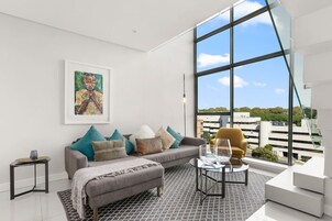 A modern living room with a grey sofa, a glass coffee table, and a large window offering a city view.