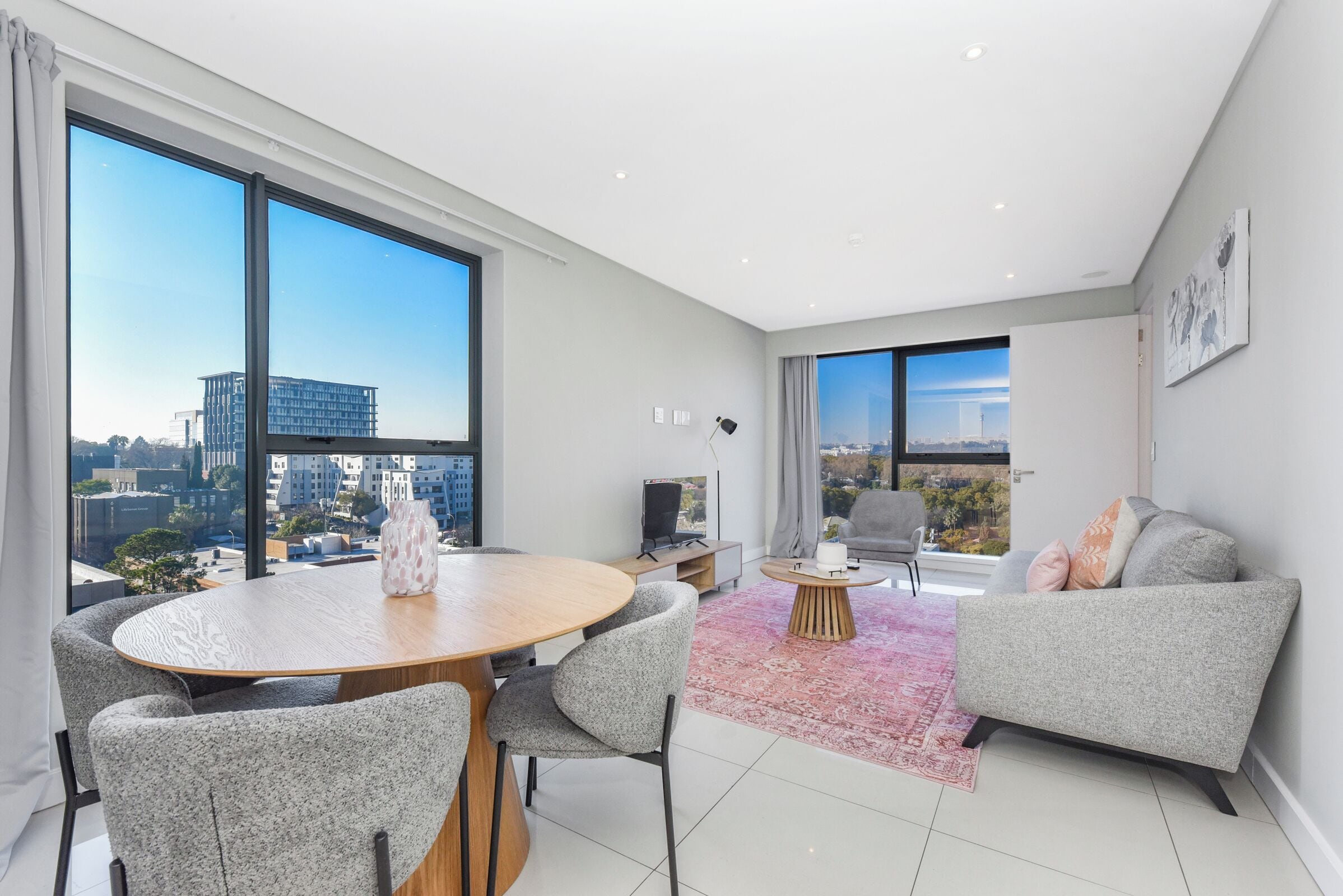 A modern living room with a dining table, grey upholstered chairs, a grey sofa, and large windows offering city views.