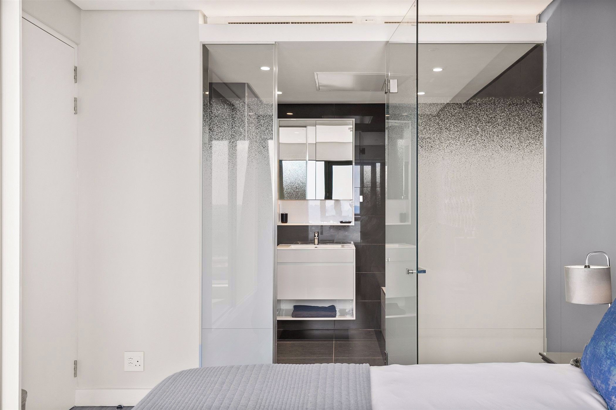 A modern bedroom with a bed, a nightstand, a lamp, and a glass-enclosed bathroom with a sink and mirror.