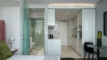 A modern hotel room with a bed, a desk, a chair, a small table, a lamp, and a kitchenette area.