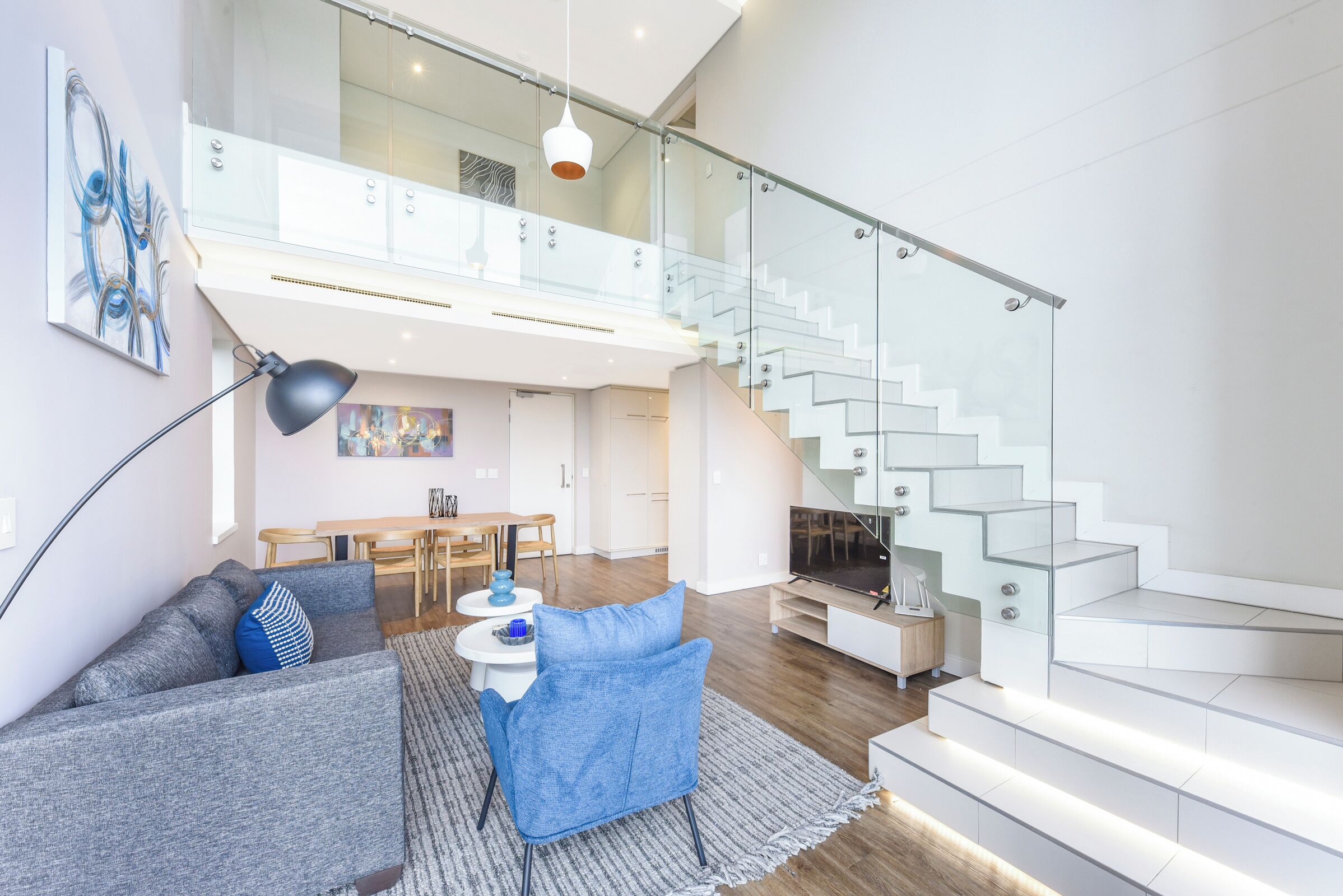 A modern living room with a sofa, a coffee table, and a TV. There is a staircase with glass railings leading to an upper level.