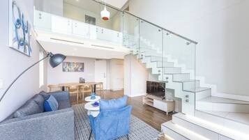 A modern living room with a sofa, a coffee table, and a TV. There is a staircase with glass railings leading to an upper level.