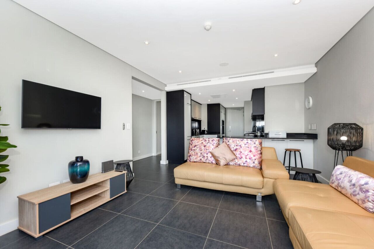 A modern living room with a flat-screen TV, a sofa, a coffee table, and a kitchen area in the background.
