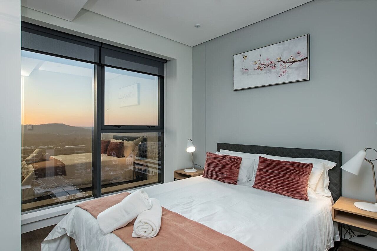 Une chambre d’hôtel moderne avec une grande fenêtre, un lit avec du linge de maison blanc, une table de chevet avec une lampe et un tableau au mur.