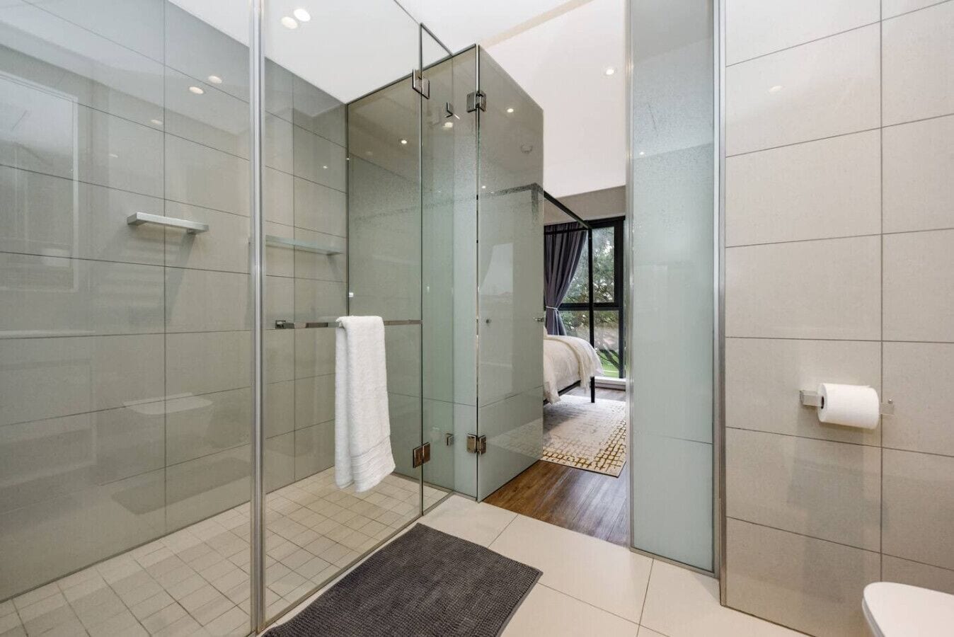 Un baño moderno con cabina de ducha de vidrio, inodoro y una habitación visible con cama y ventana.