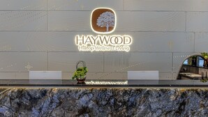 A reception desk with a logo for Haywood Hilltop Wellness Living, a potted plant, and a modern interior design. - HAYWOOD HILLTOP WELLNESS LIVING (Genting Highlands)
