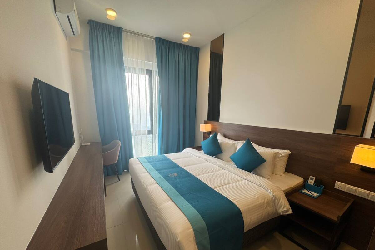 A hotel room with a large bed, a TV mounted on the wall, a desk with a lamp, and a window with curtains.
