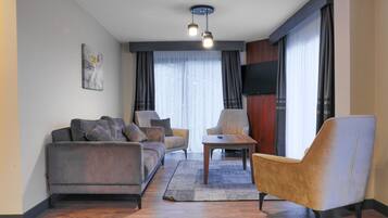 Una sala de estar moderna con sofá, sillón y mesa de centro, un televisor de pantalla plana montado en la pared y un amplio ventanal con cortinas.