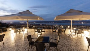 En udendørs terrasse med borde og stole under parasoller, med udsigt over by og vand.