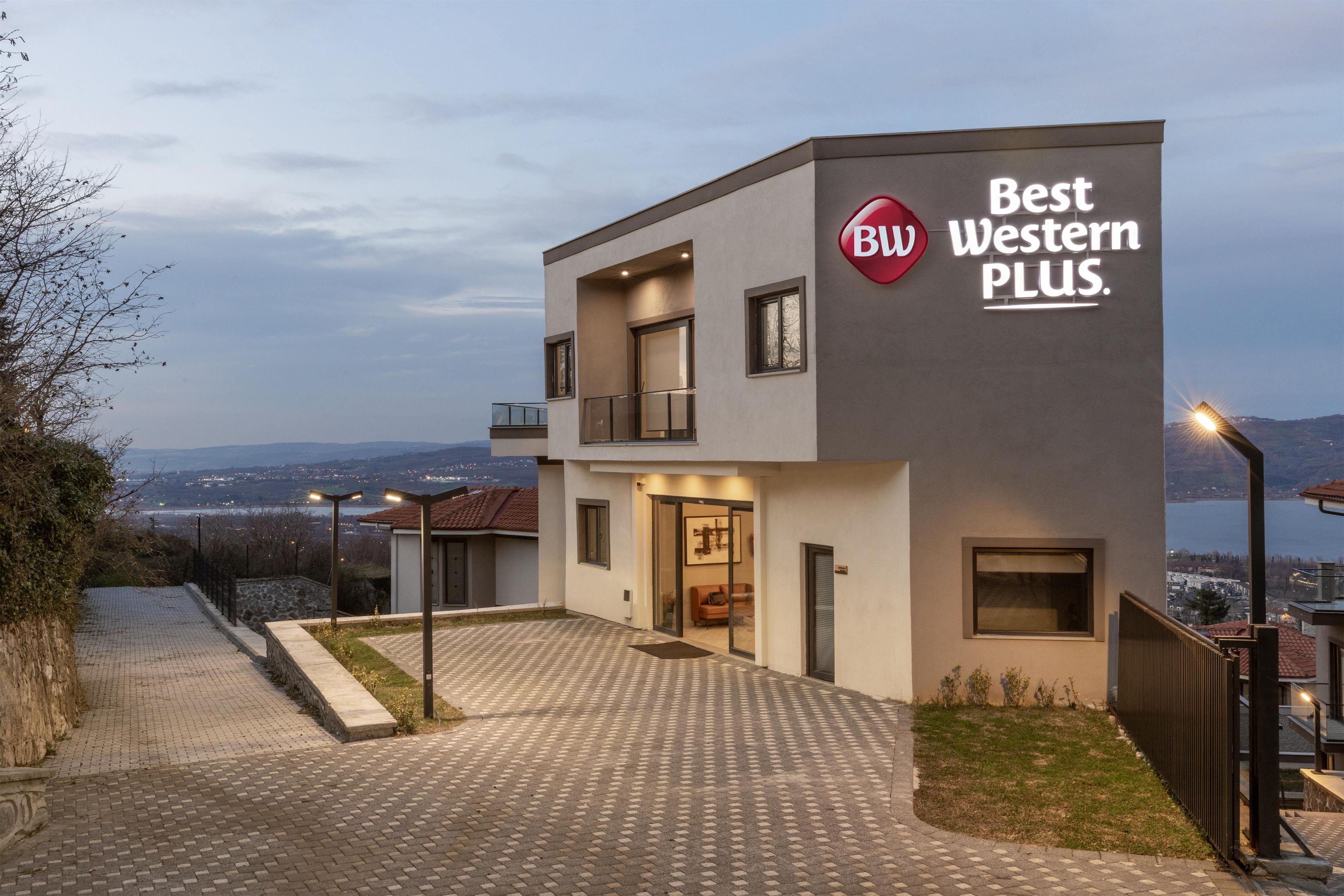 A modern building with a sign reading "Best Western PLUS." It features a paved entrance, a balcony, and a view of a body of water in the background.