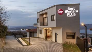 A modern building with a sign reading "Best Western PLUS." It features a paved entrance, a balcony, and a view of a body of water in the background.