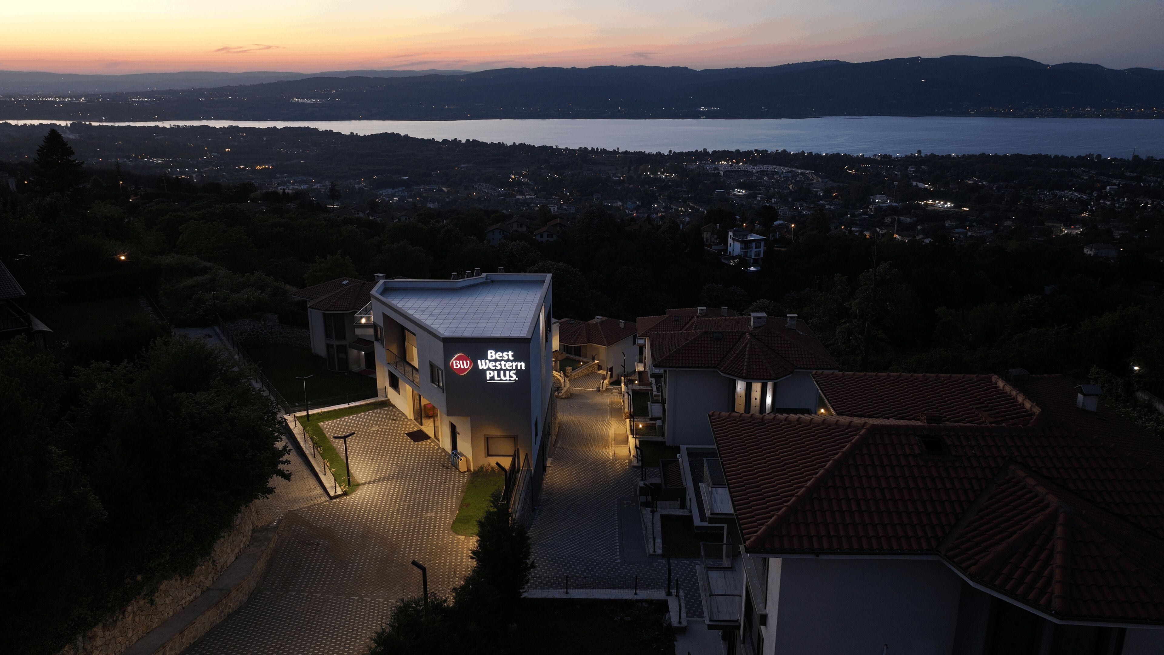 A modern building with a sign 'Best Western PLUS' at dusk, surrounded by residential buildings and a lake in the background.