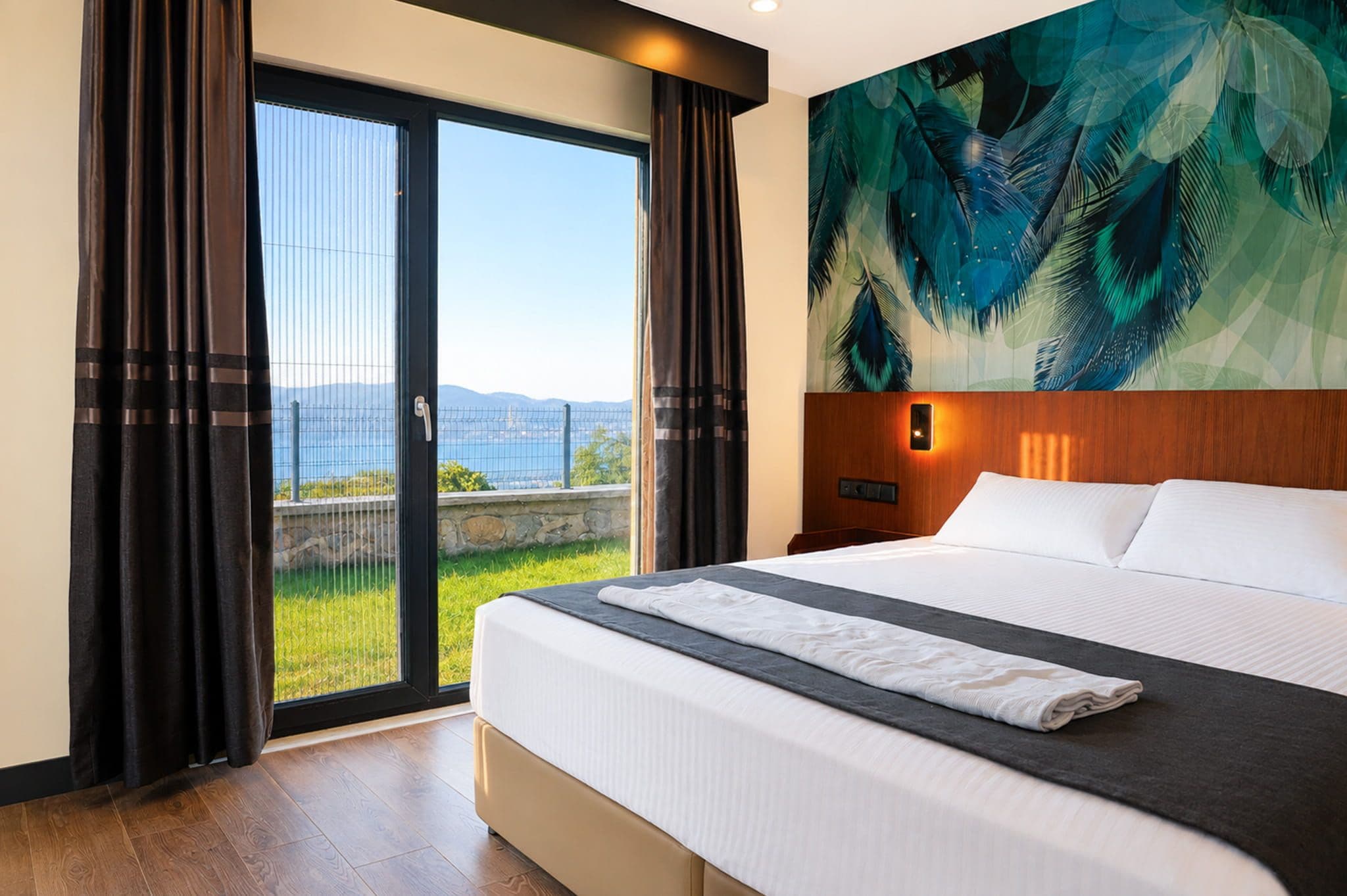 A hotel room with a large bed, a view of the sea through a glass door, and a tropical-themed wall mural.