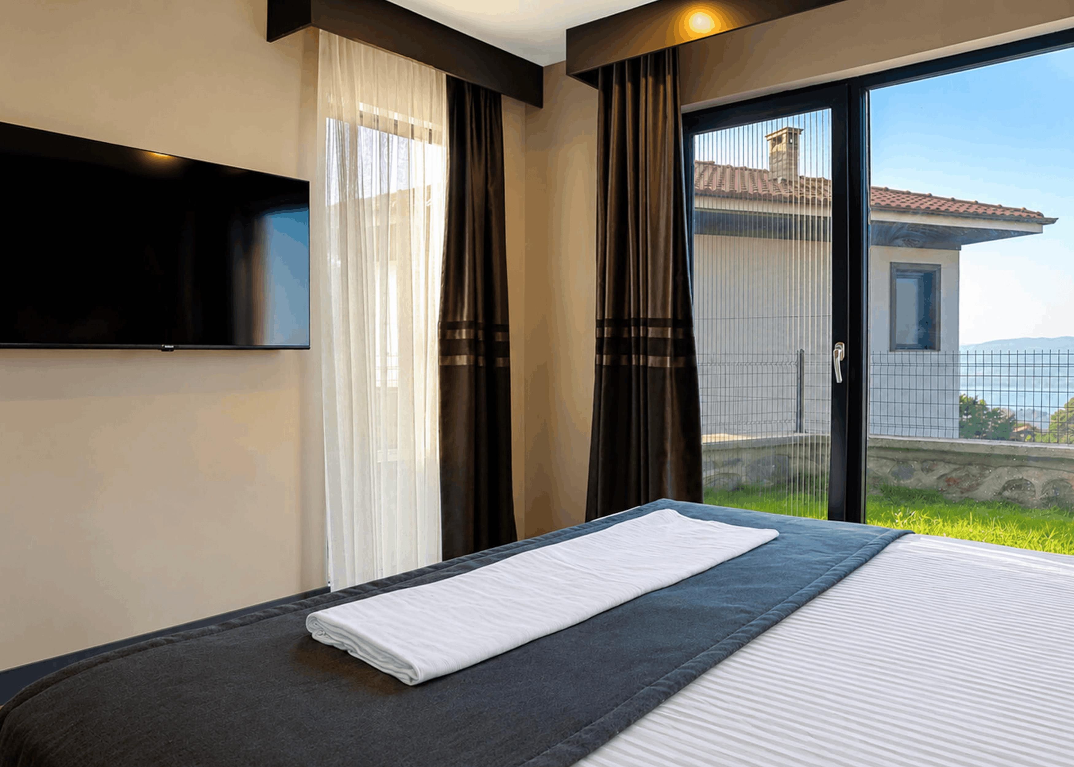 A hotel room with a bed, a flat-screen TV mounted on the wall, and a sliding glass door leading to a balcony with a view of a building and greenery.