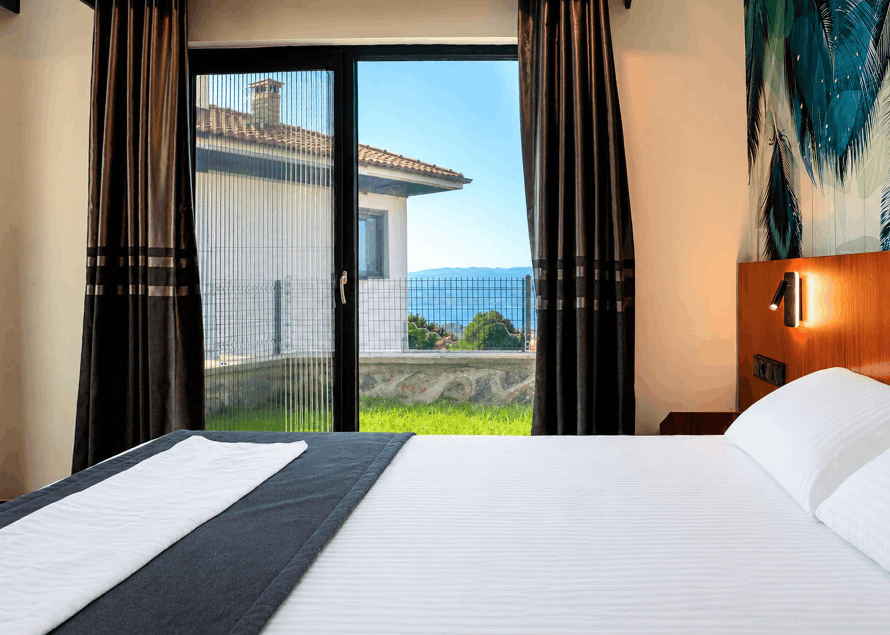 Ein Hotelzimmer mit Bett, Blick auf ein Gebäude durch eine Glastür und einem Nachttisch mit Lampe.