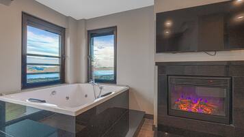 A modern bathroom with a large bathtub, two windows with scenic views, and a fireplace.