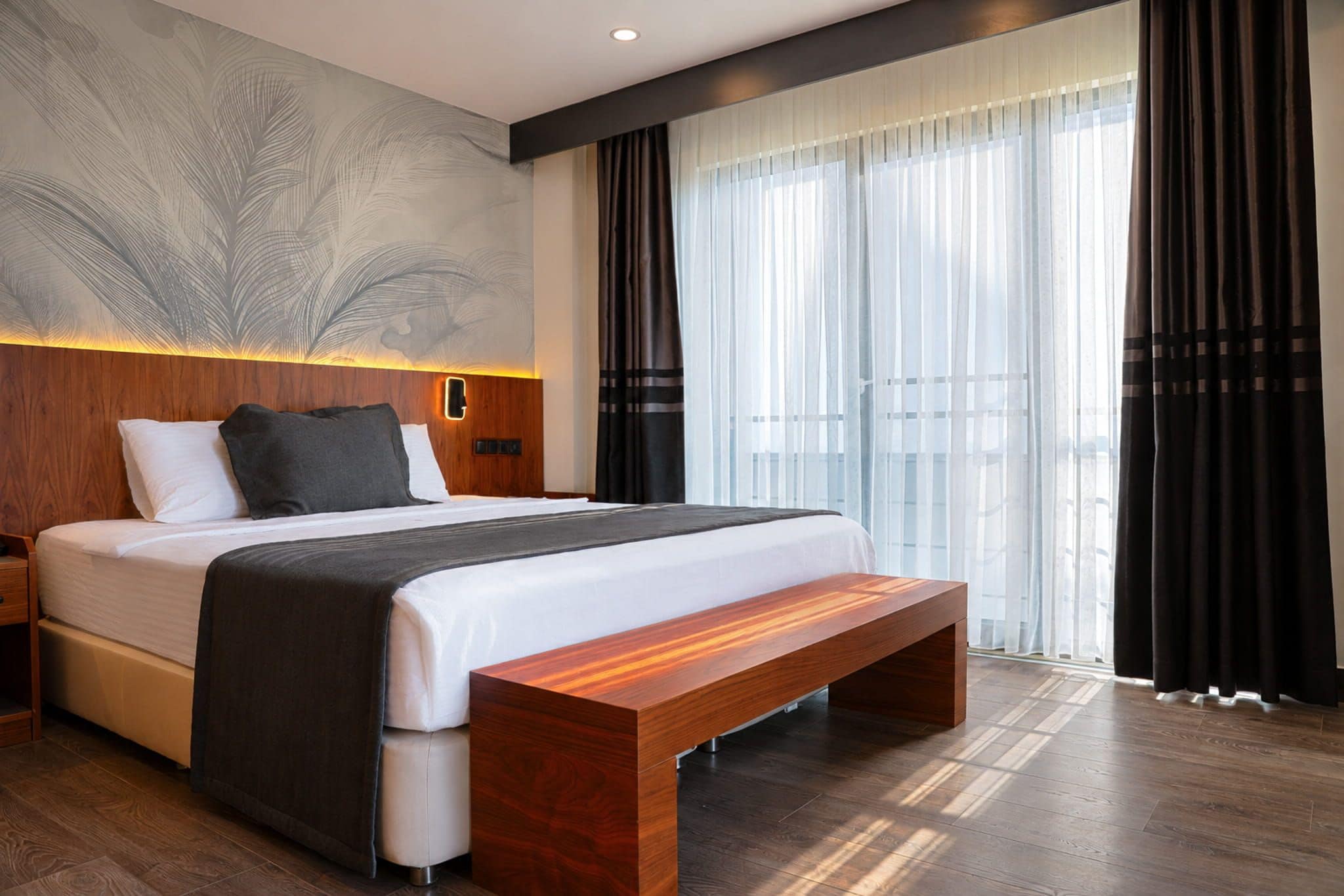 A modern hotel room with a large bed, a wooden bench, a wall-mounted light, and sheer curtains.