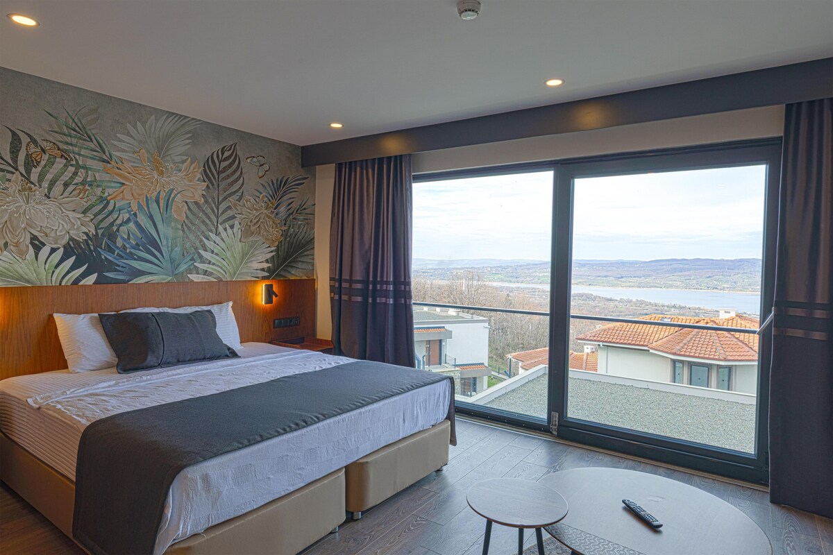 A hotel room with a large bed, a small table, a view of the city through a large window, and a wall mural.