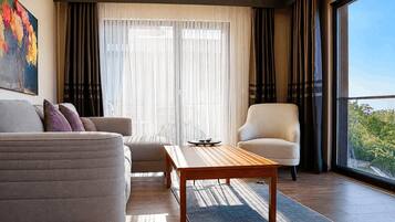 A modern living room with a sofa, armchair, and coffee table, large windows with curtains, and a view of greenery outside.