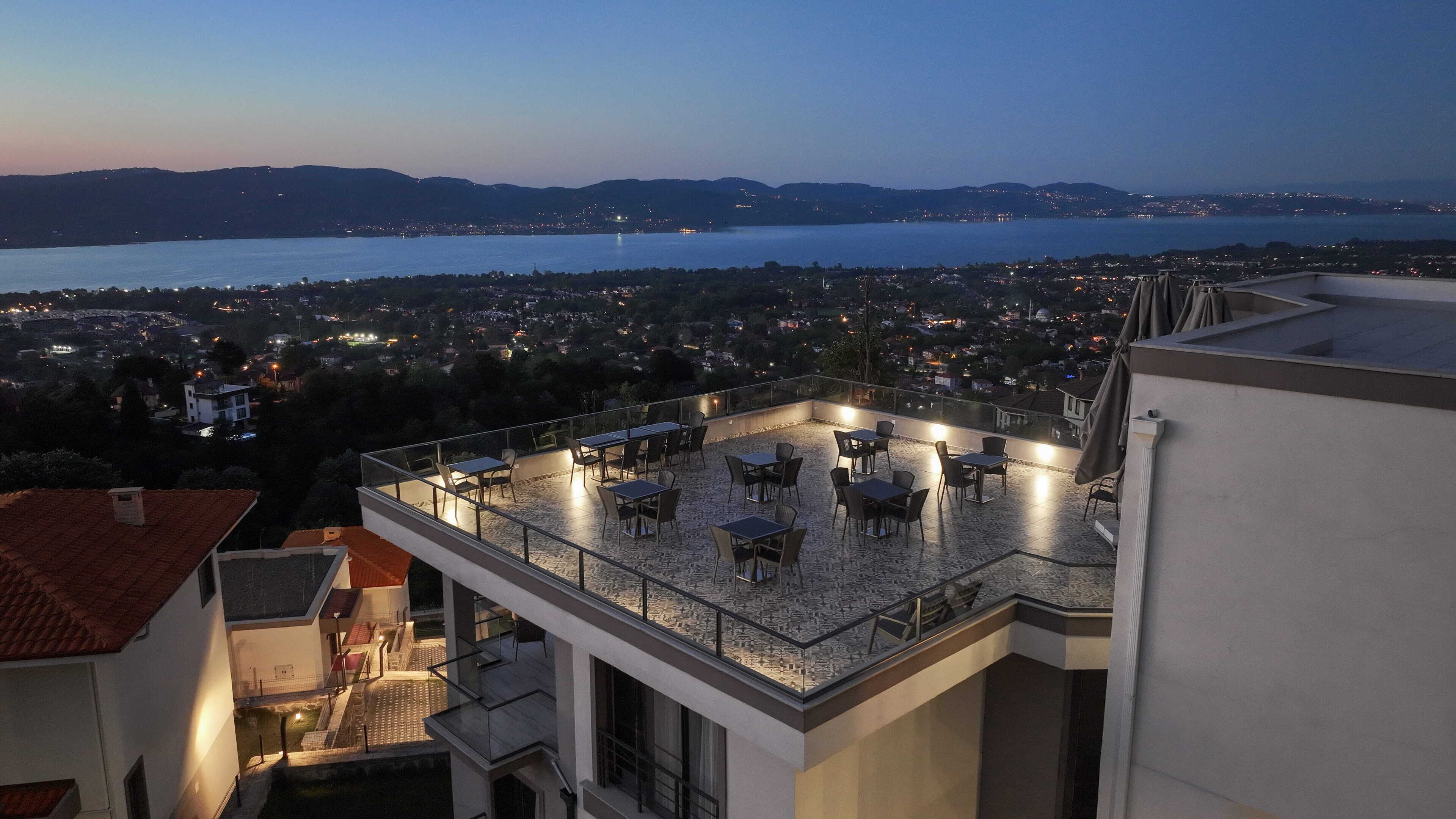 A rooftop terrace with tables and chairs, overlooking a cityscape and a body of water.