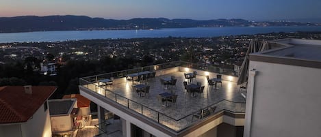 Terraza en la azotea con mesas y sillas, con vistas a un paisaje urbano y una masa de agua.