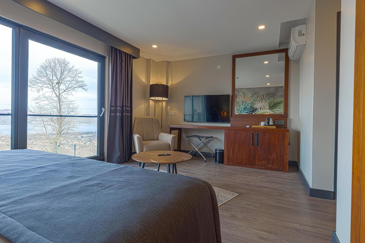 A hotel room with a large bed, a desk with a chair, a TV, and a view of the city through a large window.