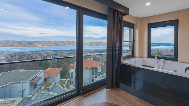 Et moderne badeværelse med et stort badekar, udsigt til en sø og en balkon med udsigt over et boligområde.