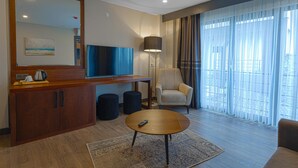 A modern hotel room with a wooden cabinet, a round coffee table, a sofa, a chair, a lamp, a TV, and a large window with curtains. - Best Western Plus Sapanca (Sapanca)