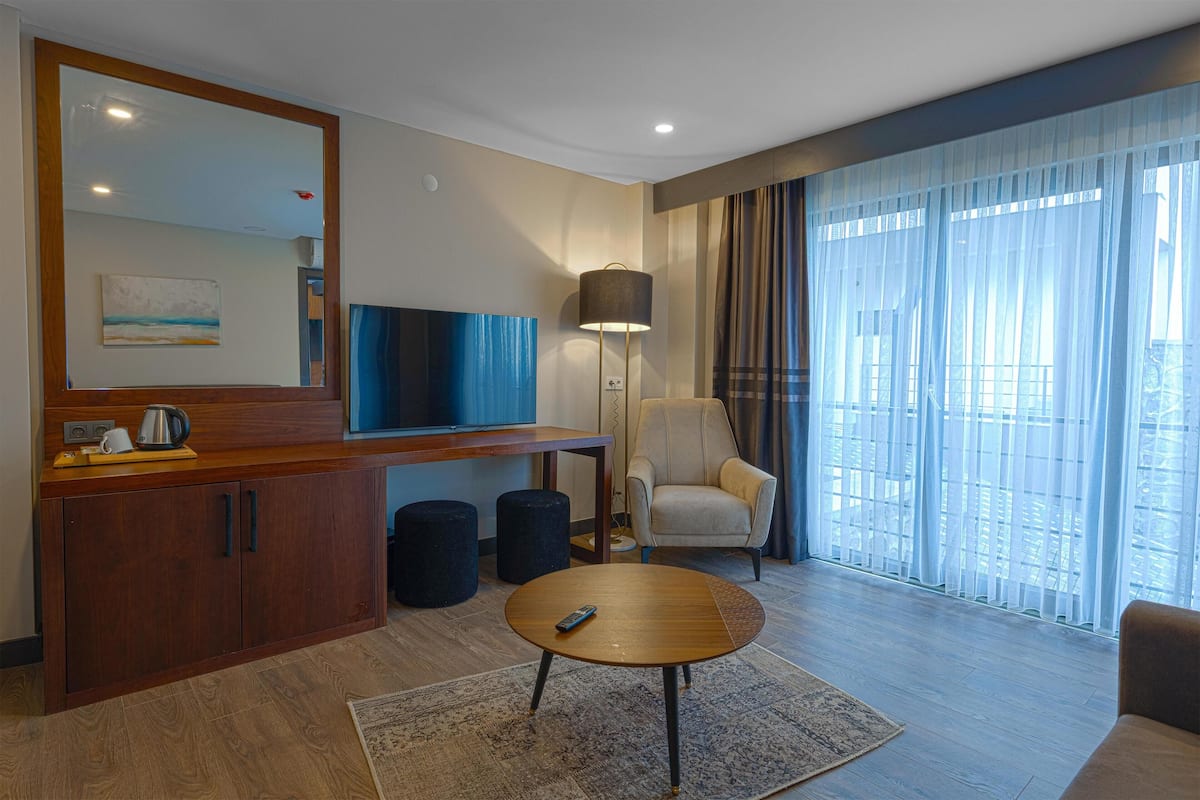A modern hotel room with a wooden cabinet, a round coffee table, a sofa, a chair, a lamp, a TV, and a large window with curtains.