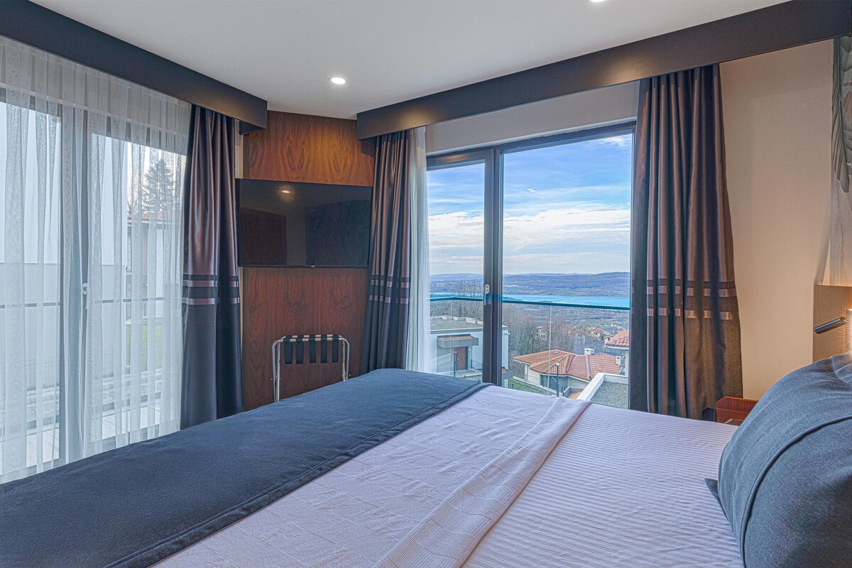 A modern hotel room with a large bed, a view of the city, and a wooden panel wall.