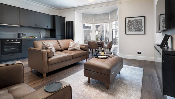 A modern living room with a brown leather sofa, a matching ottoman, and a dining area with a table and chairs. - Lake Road Apartments (Bowness-on-Windermere)