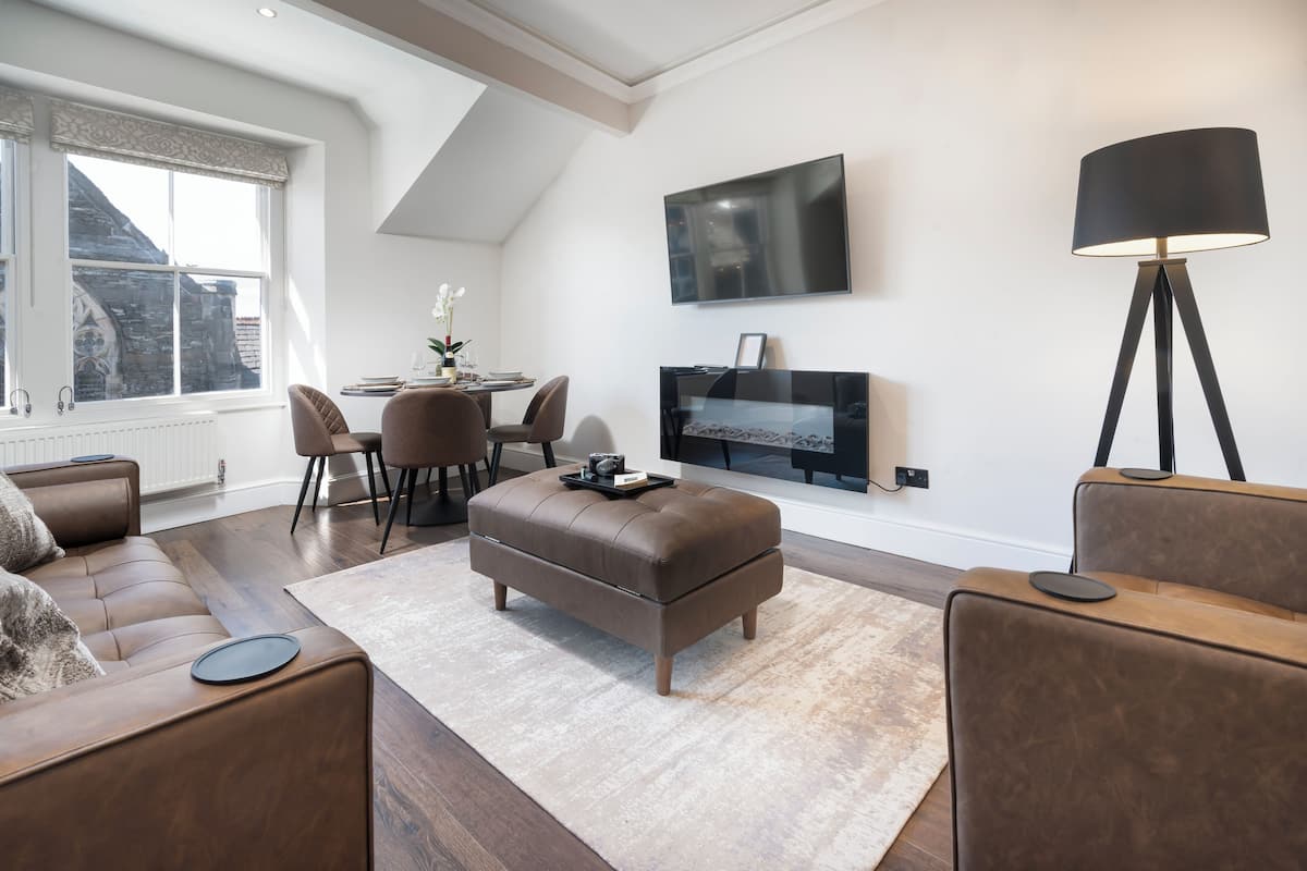 A modern living room with a brown leather sofa, a matching ottoman, a dining area with a table and chairs, a flat-screen TV mounted on the wall, and a floor lamp.