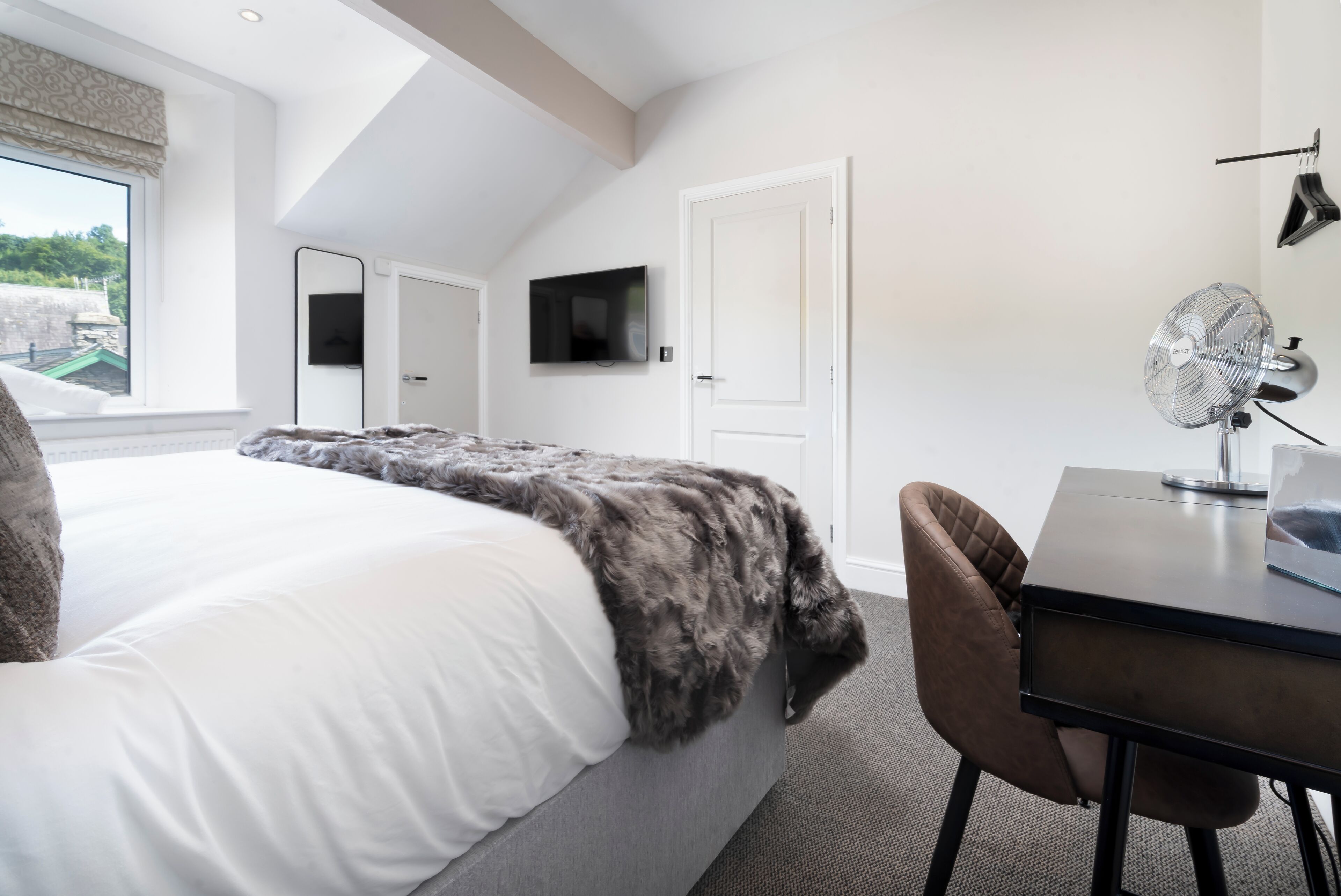 A bedroom with a bed, a desk, a chair, a TV, and a fan.