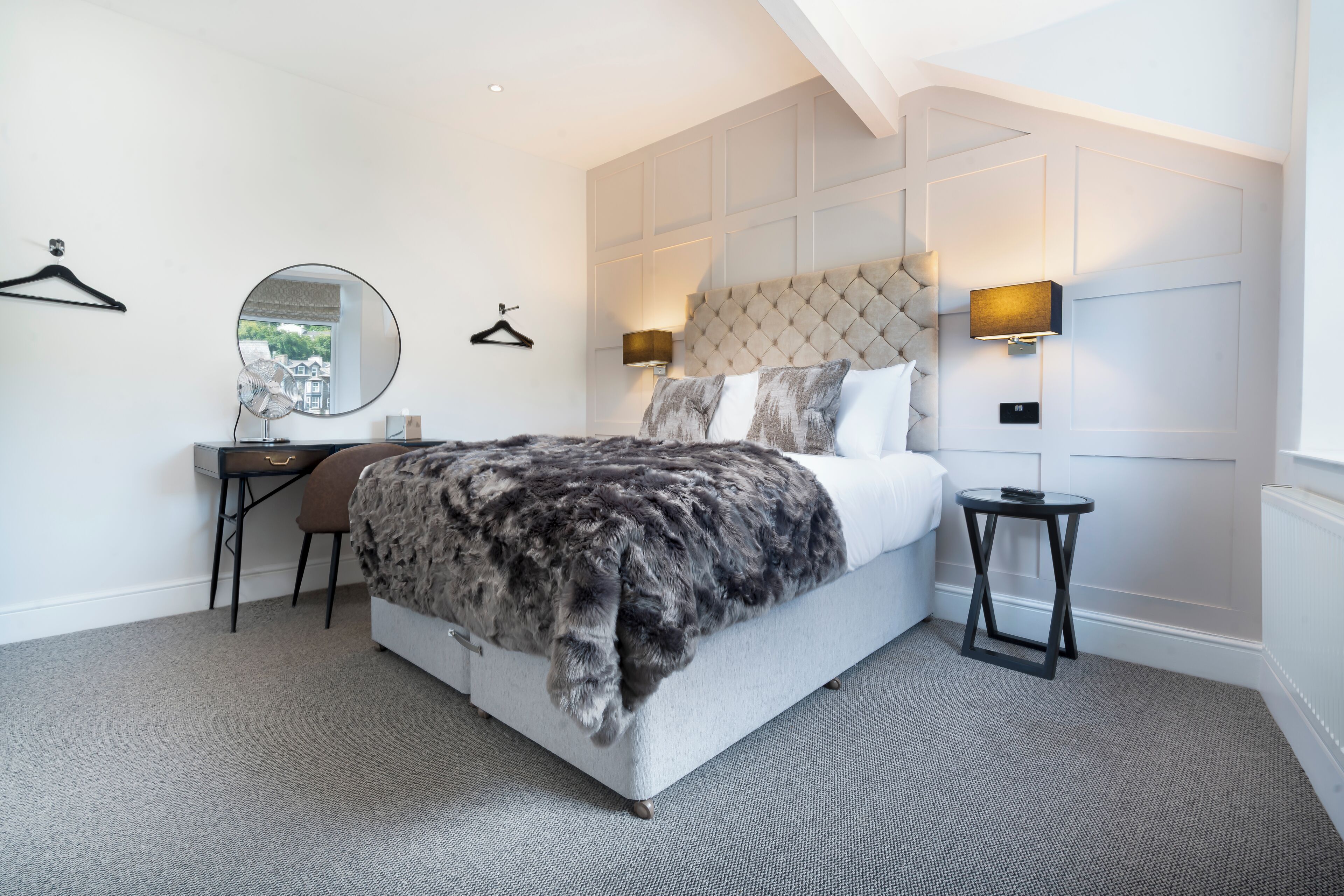 A modern bedroom with a tufted headboard, a round mirror, a bedside table with a lamp, and a large bed with a fur throw.