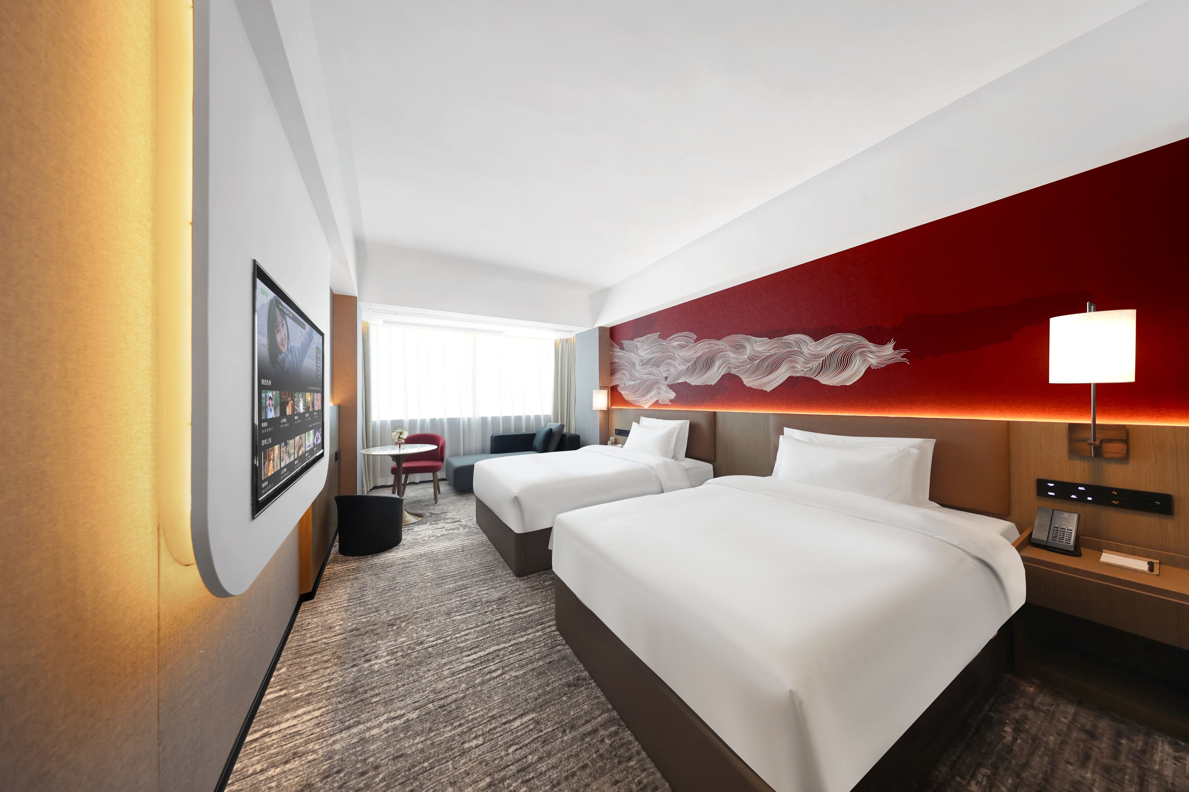 A hotel room with two beds, a desk, a chair, a wall-mounted TV, and a red accent wall.
