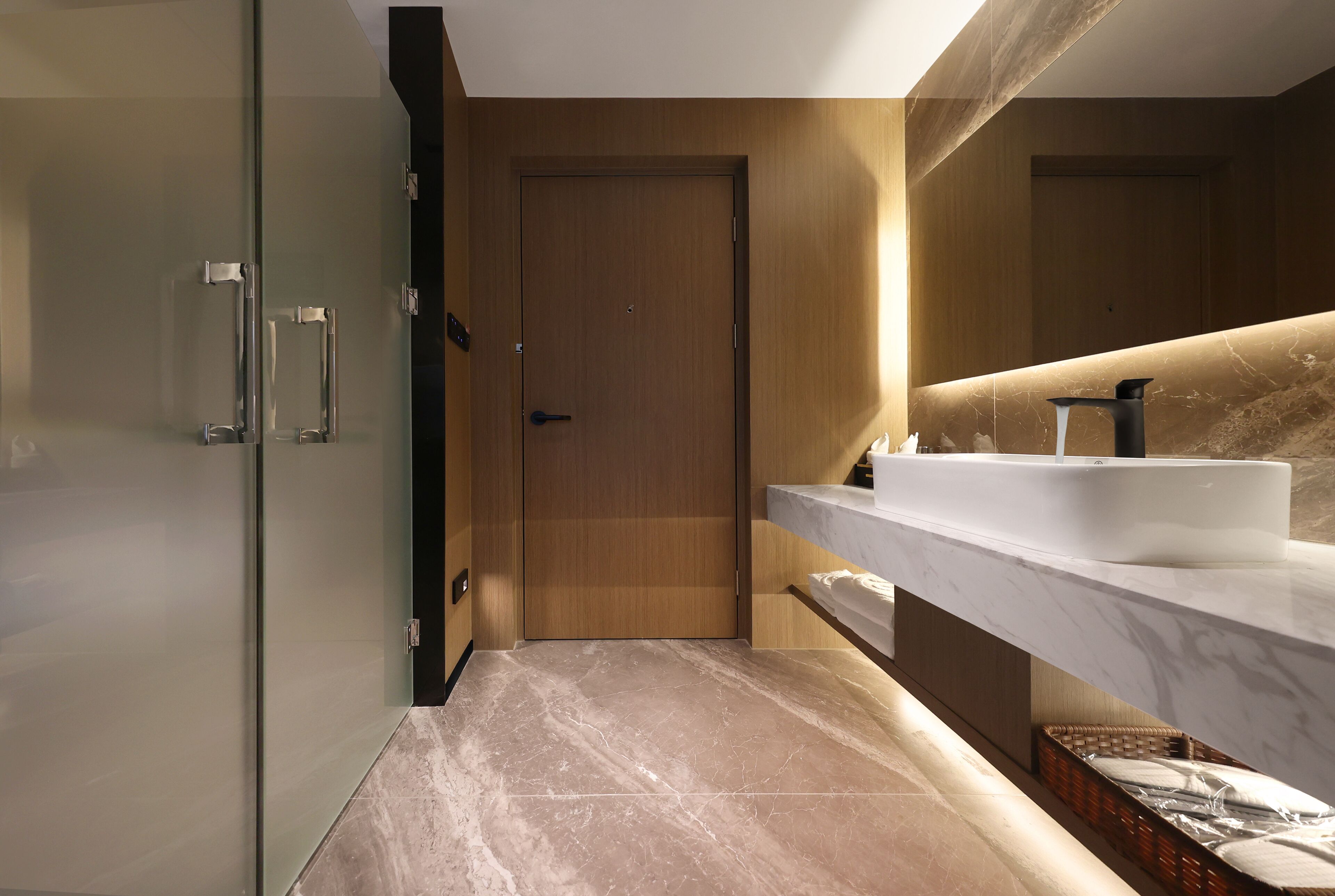 A modern bathroom with a glass shower enclosure, a wooden door, a marble countertop, and a large mirror.