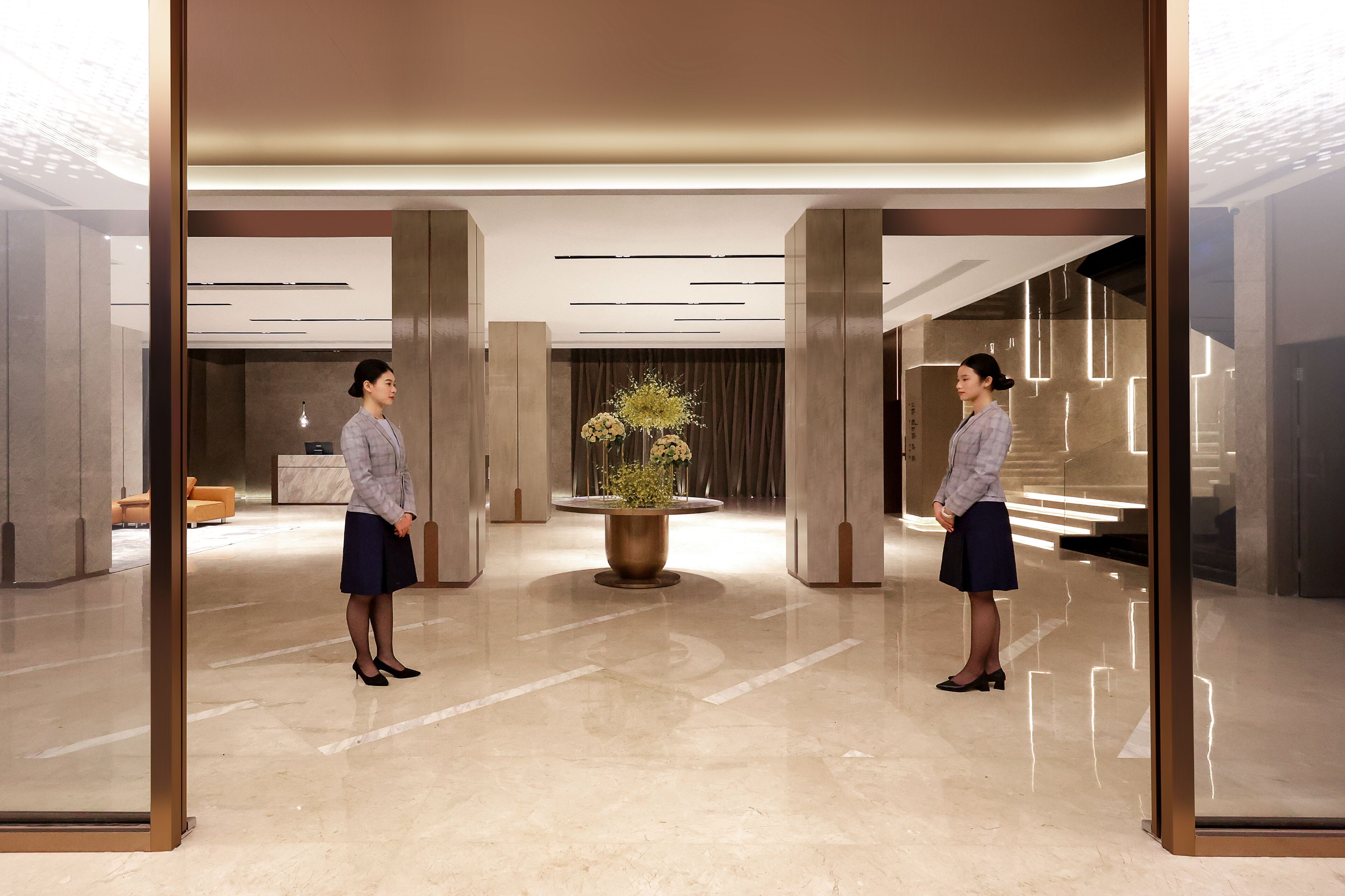 A modern lobby with a reflective floor, a central plant, and a seating area in the background.