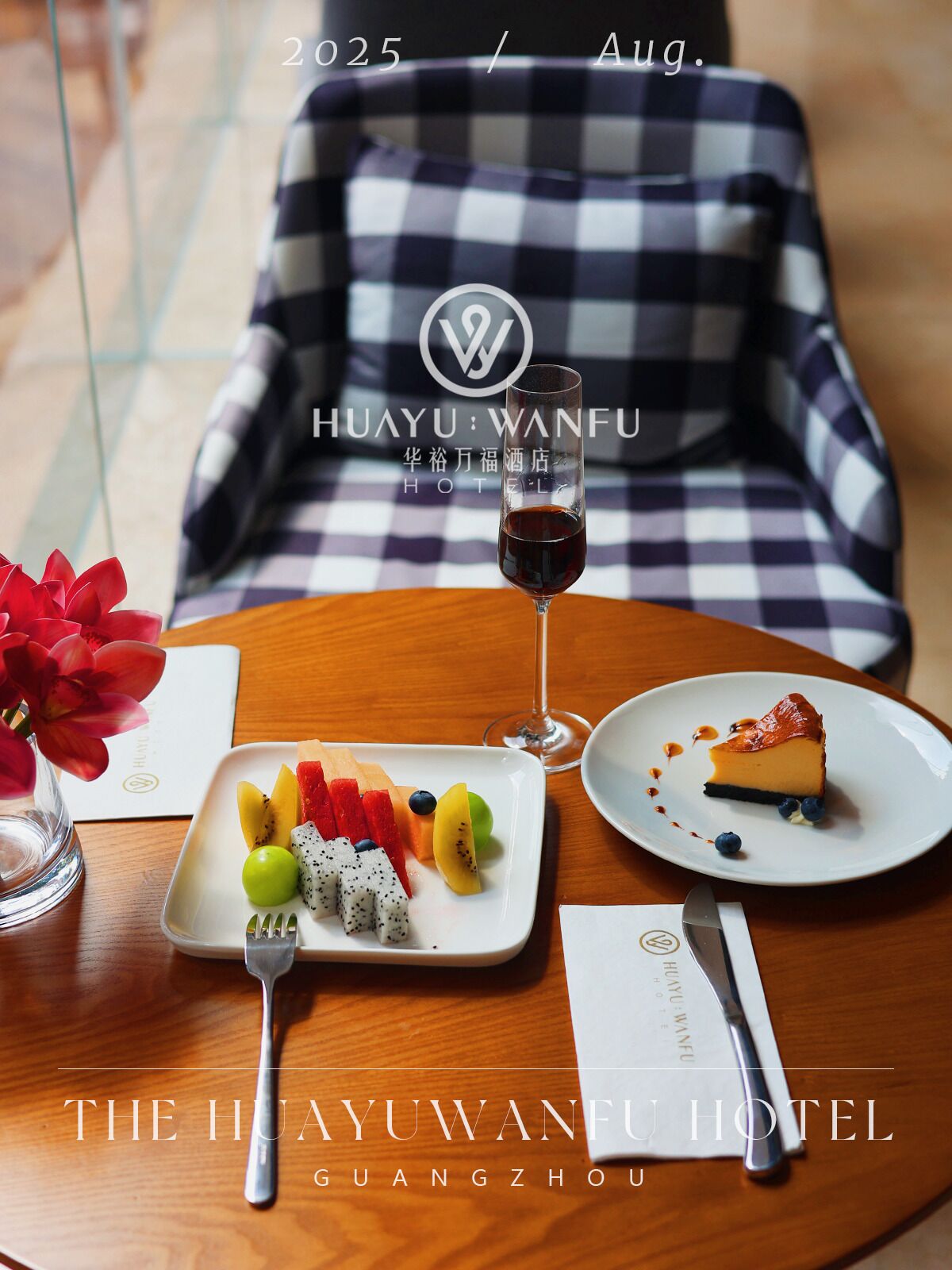 A dining table with a fruit platter, a glass of red wine, and a slice of cheesecake. There is a checkered sofa in the background and a vase with red flowers. The text 'HUAYU:WANFU HOTEL' is visible.