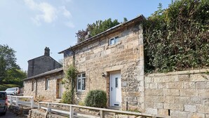A stone building with a white door and windows, a small porch, and a stone wall with vegetation. - Host Stay Brackenrigg Cottage (Morpeth)