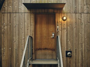 A wooden door with a metal handle and a keypad lock, a small light fixture above it, and a metal stairway leading up to the door. - SANU 2nd Home Nasu 1st (Nasu)