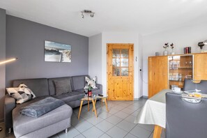 A living room with a grey sofa, a glass coffee table, a wooden door, and a dining area with a table and chairs.