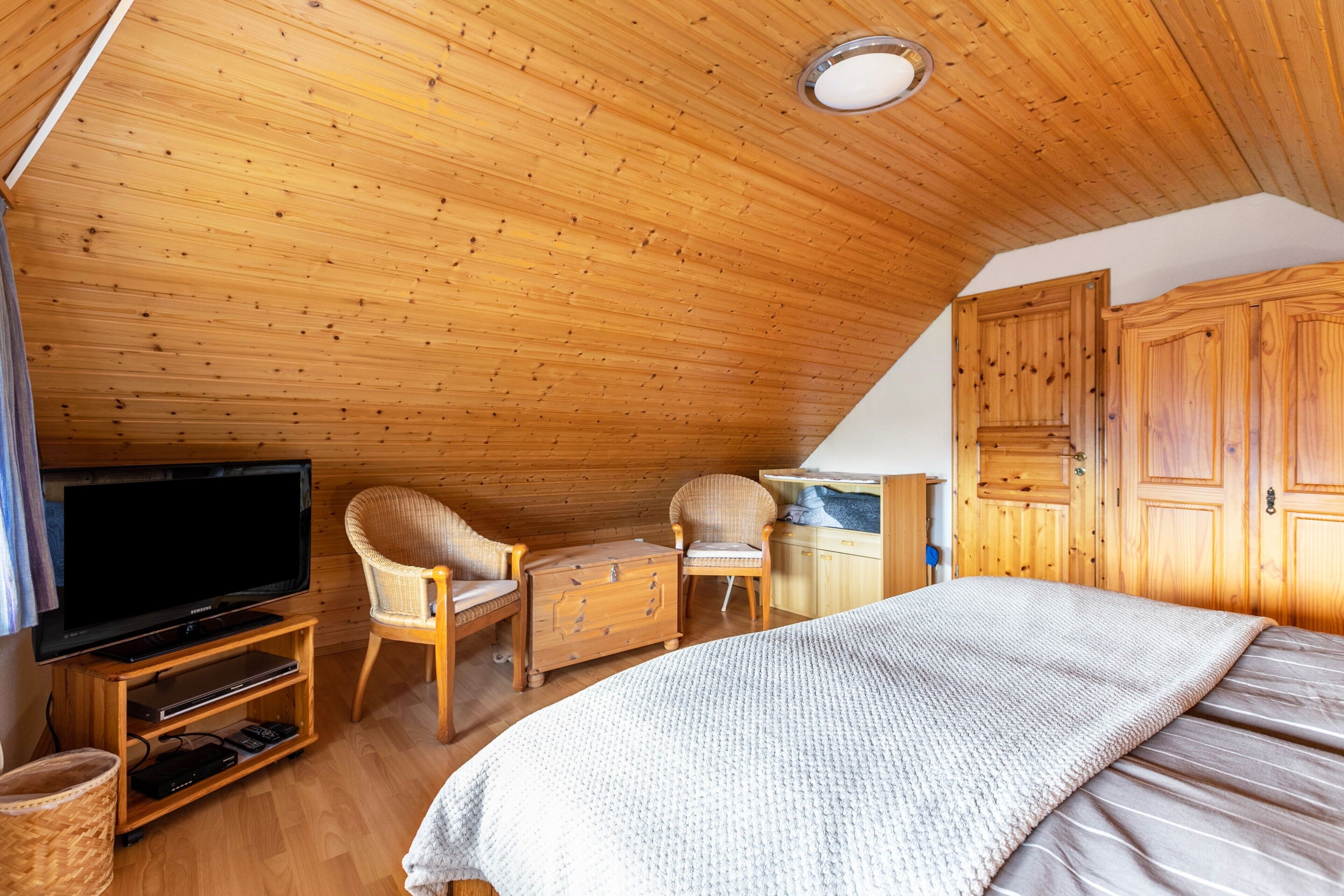 A cozy room with a wooden ceiling, a television on a wooden stand, a bed with a white blanket, a wicker chair, and wooden cabinets.