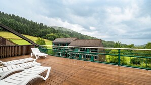 A wooden deck with lounge chairs, a green railing, and a view of a building and forested hills.