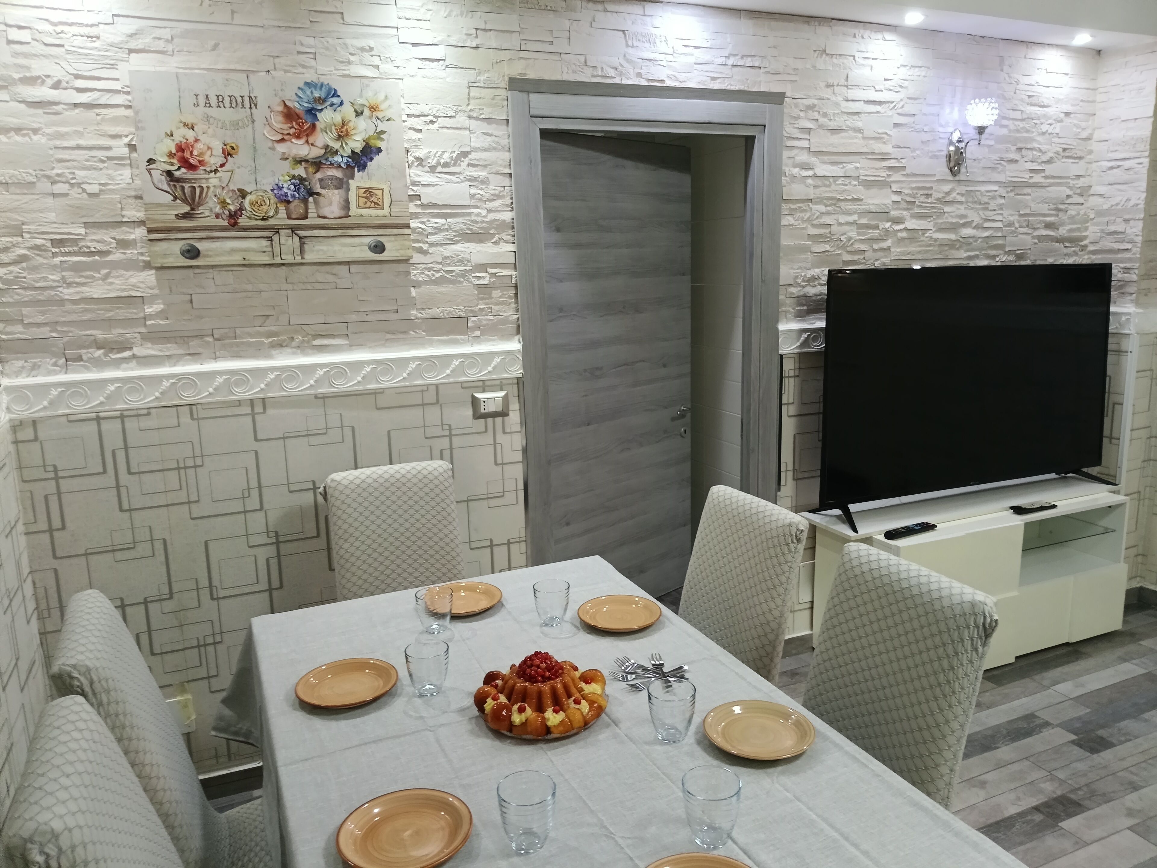 A dining area with a table set for four, a TV on a stand, and a decorative wall with a 'JARDIN' sign.