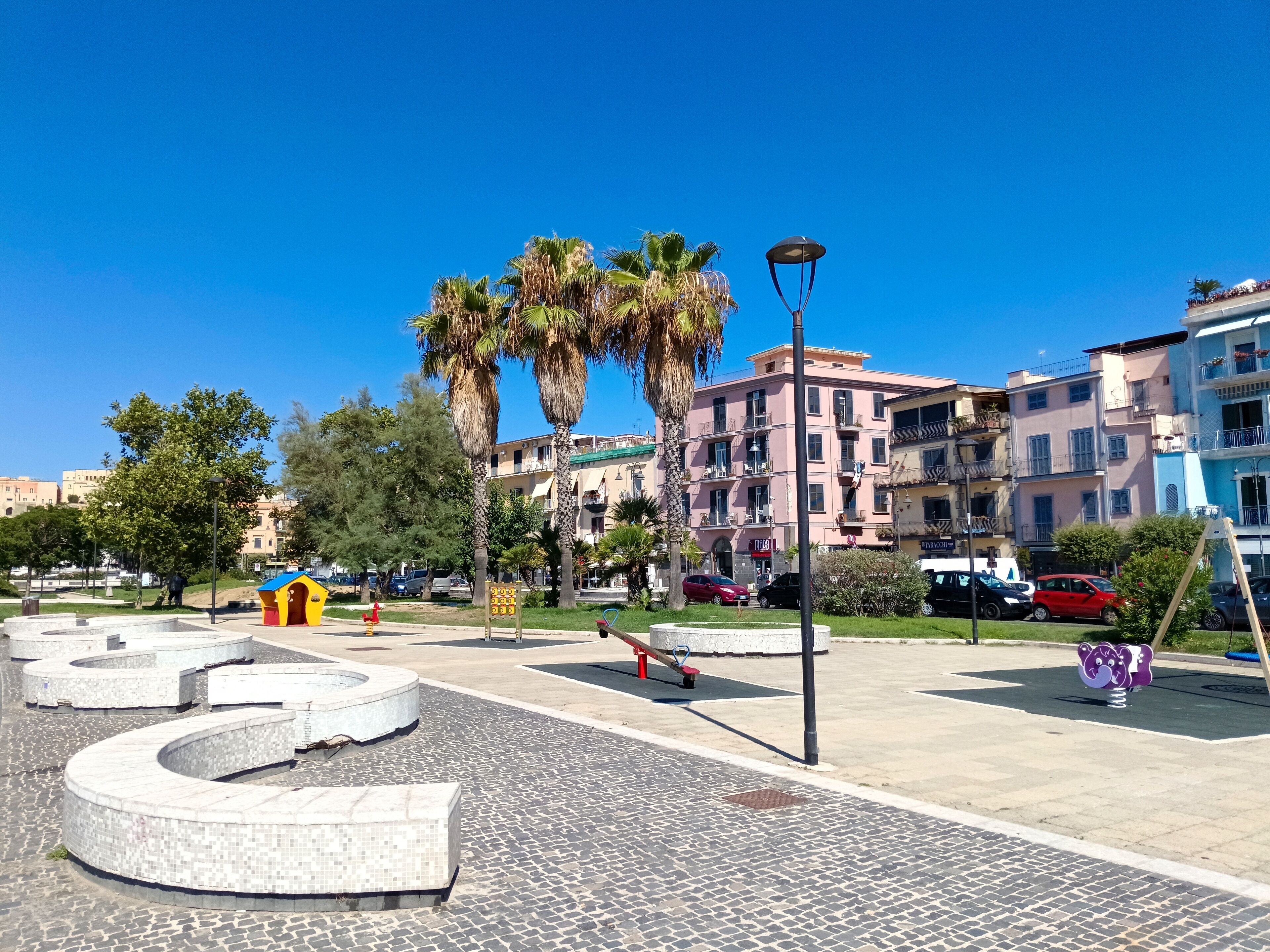 A paved plaza with a circular seating area, palm trees, and multi-story buildings in the background.