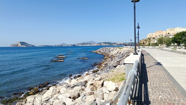 Un passeig marítim amb un costanera rocosa, mar blau clara i una vista llunyana d'un turó amb edificis.