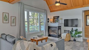 Una sala de estar acogedora con chimenea, un sofá, un televisor y ventanas amplias con vistas al exterior.